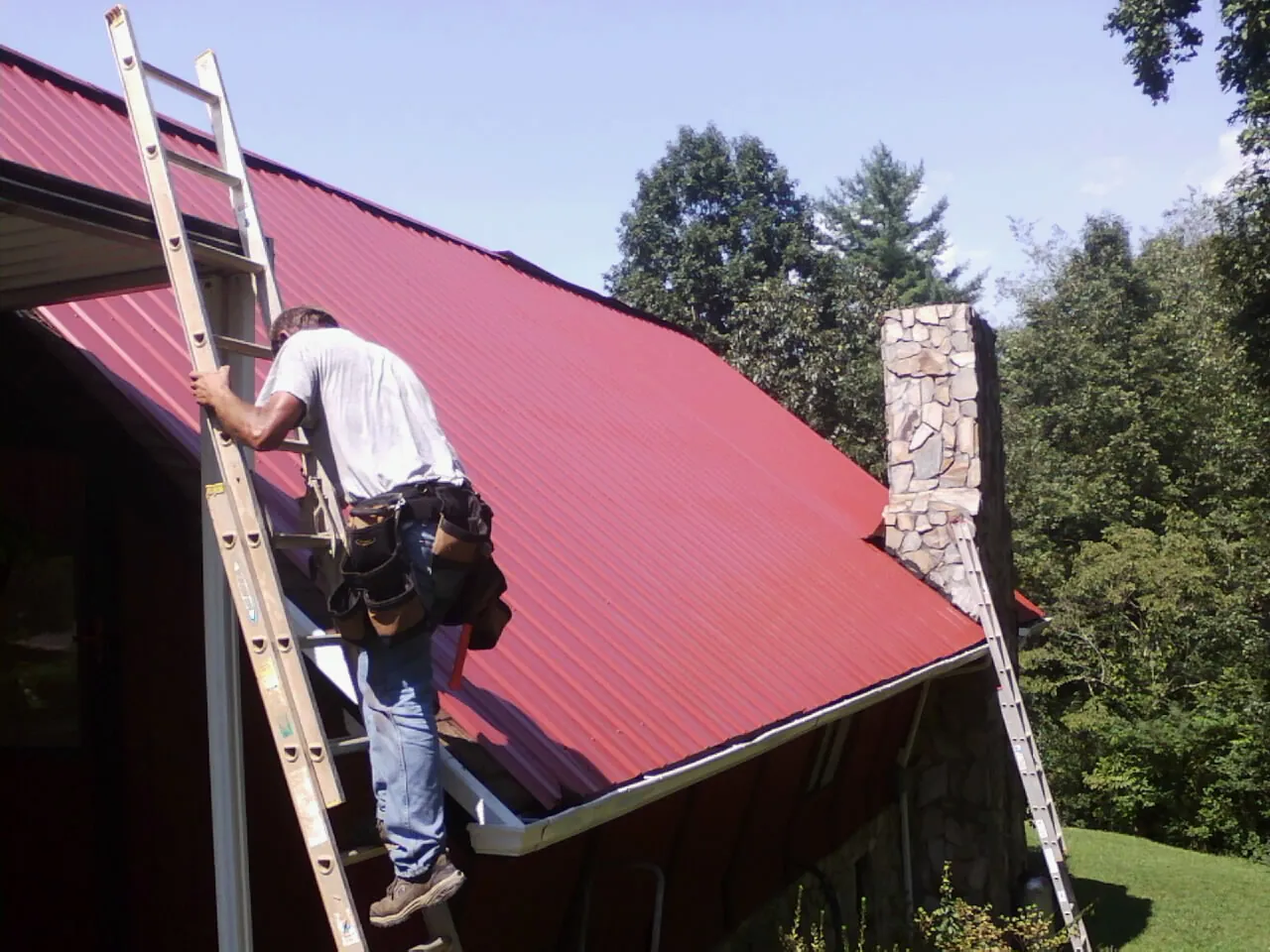 Roofer on a ladder beside a red metal roof and stone chimney — Soffit & Fascia Repair by Roofing Brothers in East Providence